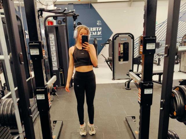 VioletaNova, con piccoli seni, indossa un reggiseno sportivo nero, leggings neri e scarpe da ginnastica bianche, e sta scattando un selfie allo specchio in palestra. I suoi capelli biondi sono raccolti in una coda di cavallo..