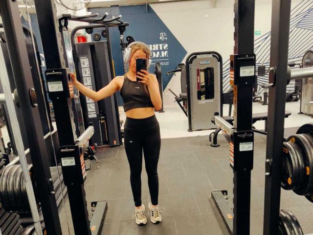 VioletaNova, avec des cheveux blonds, un débardeur de sport noir et des leggings noirs, se tient dans une salle de sport, tenant un téléphone, autoportrait devant un miroir, touchant une machine, baskets blanches, fesses visibles..
