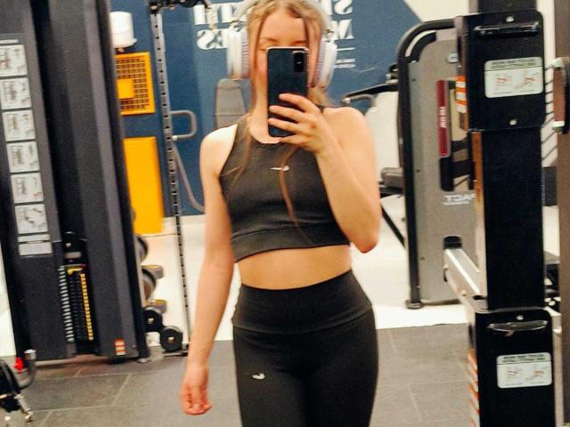 VioletaNova, avec de longs cheveux blonds, portant un débardeur de sport noir et un legging noir taille haute, tenant un téléphone dans un selfie devant un miroir de salle de sport, montrant son corps mince et plat..