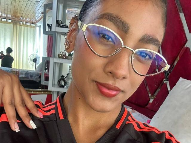 NatalyBlonder, con occhiali blu riflettenti, rossetto rosso e una maglietta sportiva nera con strisce rosse, mostra le sue unghie curate e un sorriso sicuro. La sua orecchio è adornata da un orecchino dorato..
