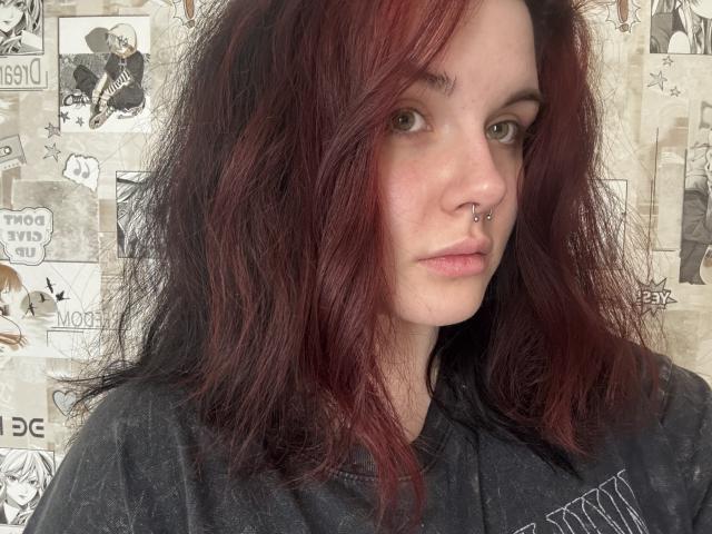 AnnaRenny, com cabelo ruivo e preto, desalinhado, com piercing no nariz, vestindo uma camisa cinza e com uma expressão séria, olhando para a câmera. Pele clara, olhos verdes e maquiagem discreta..