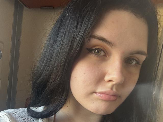 AnnaRenny, met zwart haar, bruine ogen en een kleine neusring, kijkt naar de camera, draagt een witte top, heeft een lichte huid, licht open lippen en subtiele eyeliner..