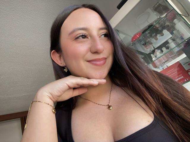 Rosalie Dupont, con cabello castaño largo, piel clara y una sonrisa sutil, apoyando el mentón en la mano, vestida con una blusa negra que deja ver parte del escote, y una pulsera dorada..