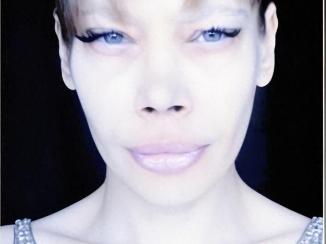 LorraineRux's face with pale skin, blue eyes, and light pink lips, wearing a pearl necklace, with short brown hair and black eyeliner, staring directly into the camera.