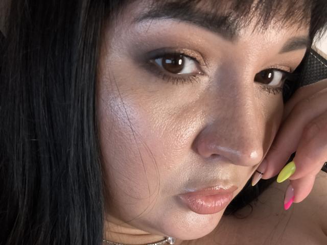 El rostro de AlinaMilf, con piel brillante, ojos marrones y cabello negro, tiene una expresión de labios fruncidos y está apoyando su uña amarilla y rosa en su mejilla..