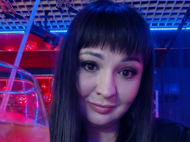 AlinaMilf, avec ses cheveux bruns foncés et lisses, sa frange et son léger sourire, tient une boisson avec une paille dans un bar éclairé au néon bleu et rouge, et montre son joli visage..
