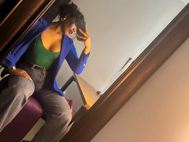 SophieSonyas, com cabelo cacheado, jaqueta azul, blusa verde e jeans, tirando uma selfie no espelho, mostrando os seios, sentada em uma cadeira vermelha, segurando um celular..