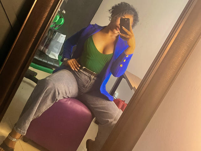SophieSonyas with curly hair, blue blazer, green ribbed top, showing big boobs, gray pants, sitting on purple bench, taking mirror selfie, holding phone, in dimly lit room.