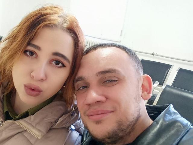 GussieBridie with orange hair and pouty lips, next to a bald, bearded dude with a smirk, both showing off cute, sexy faces, wearing jackets, in a casual selfie.