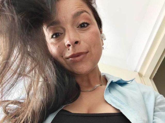 StangelarRivera a de longs cheveux bruns, une peau brun clair et de petites seins, elle porte une chemise bleu clair par-dessus un haut noir, avec un sourire discret et un collier..