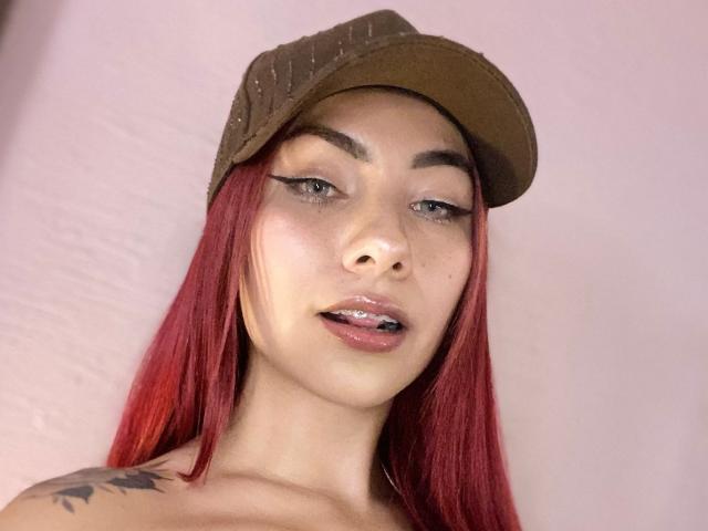 ValeryHarllow with red hair, brown cap, eyeliner, glossy lips, slight tongue out, tattoo on left shoulder, fair skin, looking at camera, soft pink lighting.