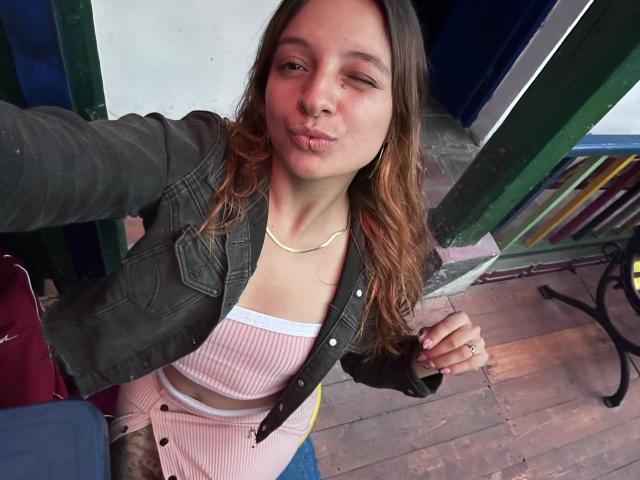 SaarahJones, labios fruncidos, guiñando un ojo, con una camiseta rosa de canalé, chaqueta negra, cabello rubio, suelo de madera, puerta verde, sosteniendo un teléfono, anillo en el dedo, informal, linda, pechos pequeños..
