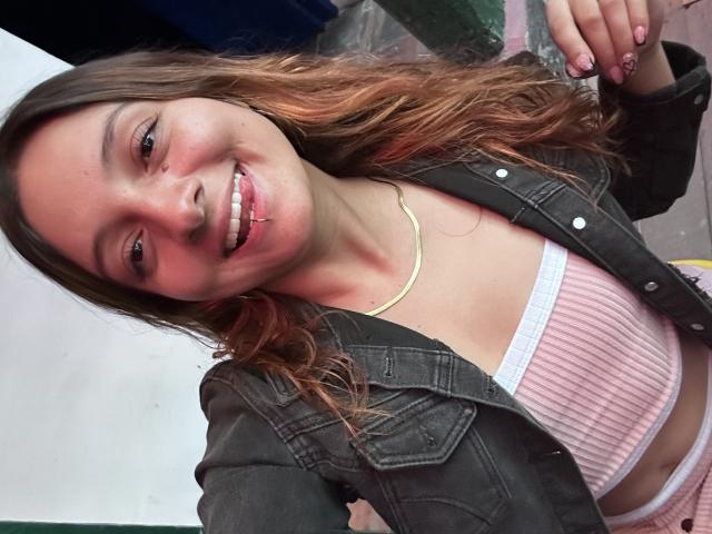 Saarah Jones, con cabello castaño ondulado, sonriendo, vestida con una chaqueta vaquera negra, una camiseta rosa de canalé y ropa interior blanca, mostrando un pequeño piercing en la nariz y un collar dorado..