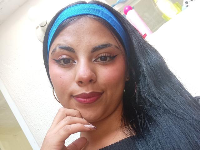 MelanySimis, com cabelo escuro, uma faixa azul na cabeça, brincos de argola e batom rosa, tem o dedo perto do queixo, exibindo seus longos olhos, realçados com cílios, e seu sorriso adorável..