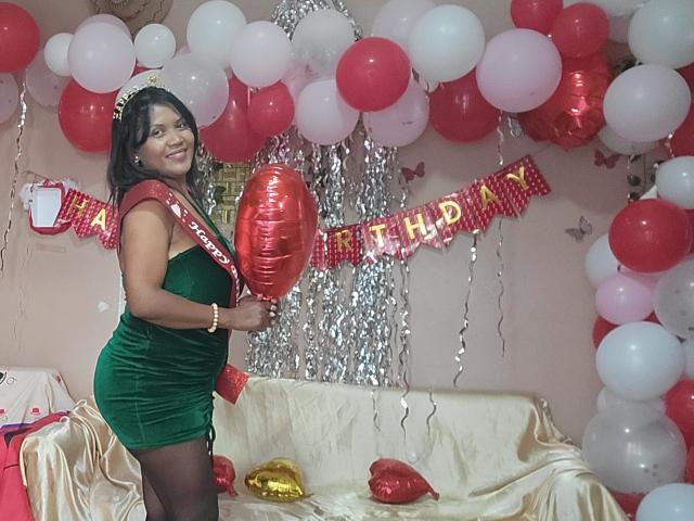 FelyLove mit dunkler Haut, schwarzen Haaren, die ein grünes Samtkleid und einen roten Gürtel trägt, einen roten Luftballon hält und in einem Raum steht, der mit roten und weißen Luftballons gefüllt ist..
