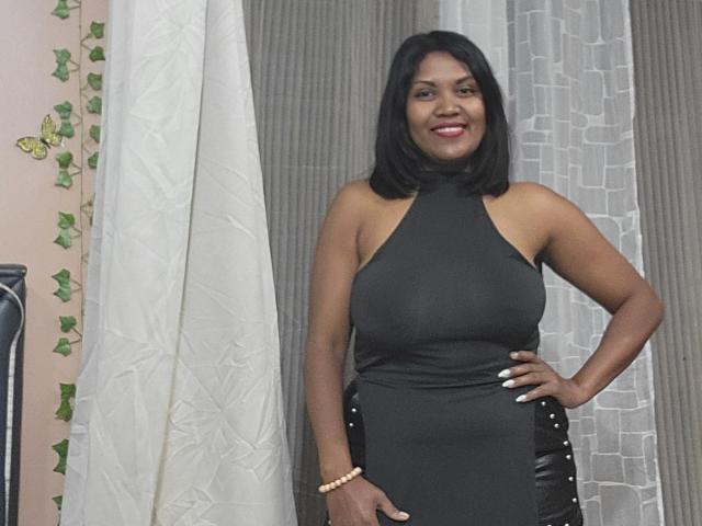FelyLove has dark skin, black shoulder-length hair, and is wearing a tight black halter dress, showing her big ass and small tits, with a smile and white nails.