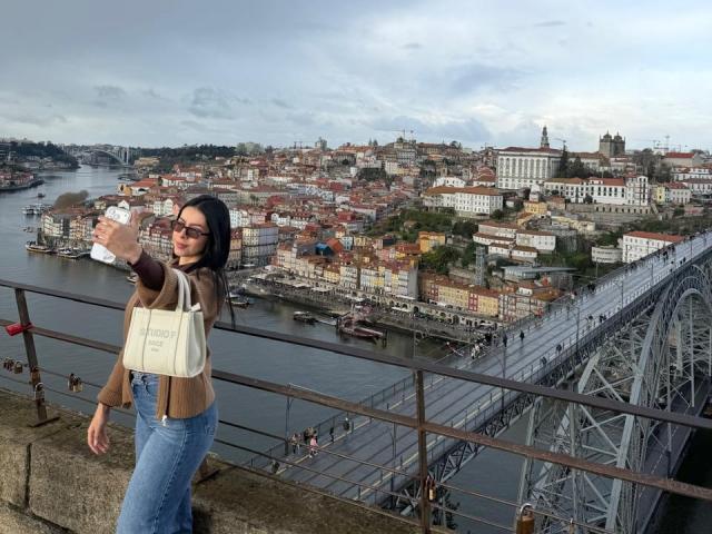 IsabellaJames with tits covered, wearing glasses, brown jacket, white bag, blue jeans, taking a selfie on a bridge, overlooking a city with a river and buildings.