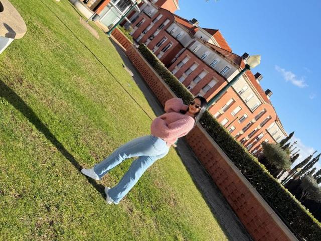 IsabellaJames stands on grass with a pink sweater, blue jeans, and white sneakers, back to camera, hands in front, looking at red brick building with green hedge.
