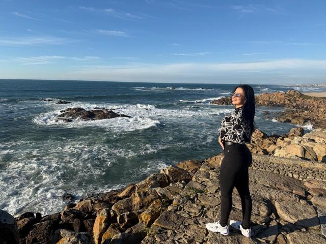IsabellaJames stands on rocky shoreline, wearing black pants and white sneakers, with long black hair, a black-and-white patterned top, and a smile, facing ocean waves.