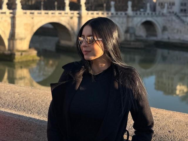 IsabellaJames with long black hair, wearing glasses and a black shirt, stands by a river with a stone bridge in the background, sunlight on her face.