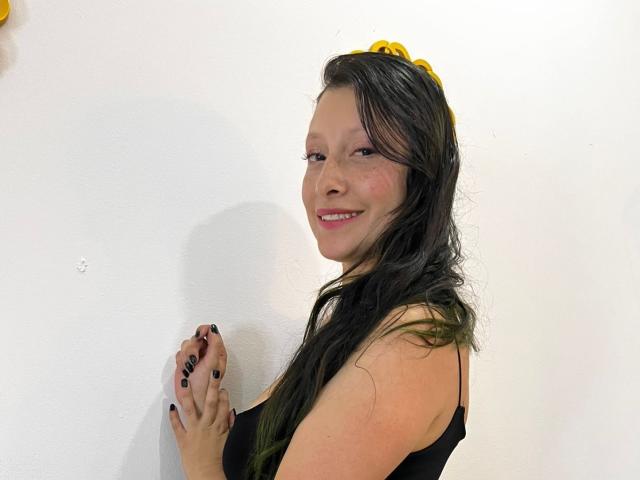 MiaSttar, con lunghi capelli neri, indossa una fascia per capelli gialla e un top nero, sorride alla fotocamera, ha le unghie nere, ed è sullo sfondo di un muro bianco. Il suo seno è visibile..