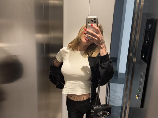 SensualMila with blonde hair, white tee, black jacket, and black pants, holding phone to her face, red nails, tattoos, black purse, in elevator, showing her tits.