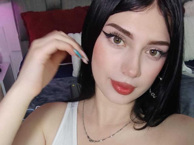 IsabellaaRossi with pale skin, dark hair, green eyes, red lipstick, white tank top, silver necklace, and blue nail polish, touching her hair, looking at the camera.