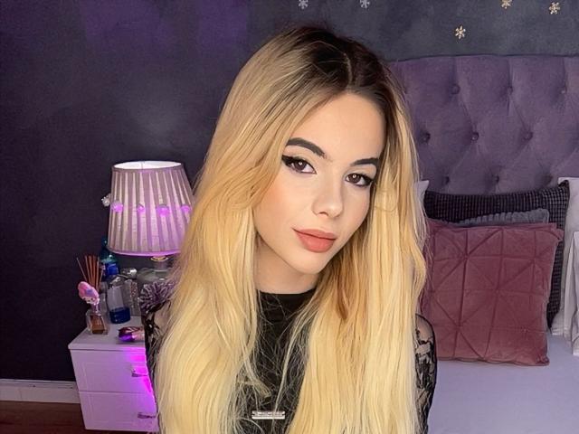 AmirraLovely with long blonde hair, full eyebrows, and pink lipstick, wearing a black lace top, standing in front of a purple-lit lamp and a tufted grey headboard.