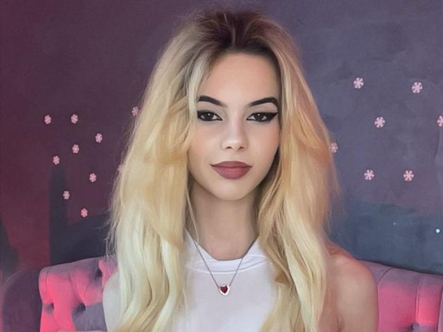 AmirraLovely has long blonde hair, dark eyebrows, and red lipstick. She's wearing a white sleeveless top with a heart necklace, and she's looking directly at the camera.