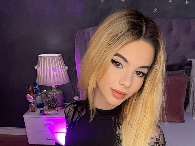 AmirraLovely with blonde hair, black lace top, and pink lipstick, looking at camera with a slight smile. Purple lamp and makeup on bedside table.