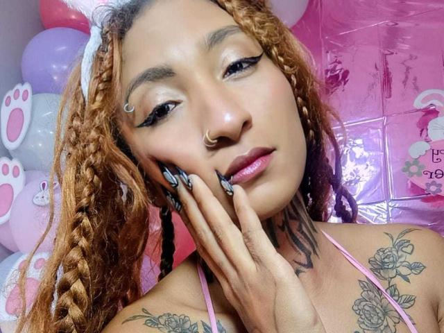 SavageKnela, com cabelo laranja trançado, piercing no nariz e tatuagens, tocando sua bochecha, mostrando lábios rosados, delineador preto e um top de alça rosa, com dedos longos pintados de preto..