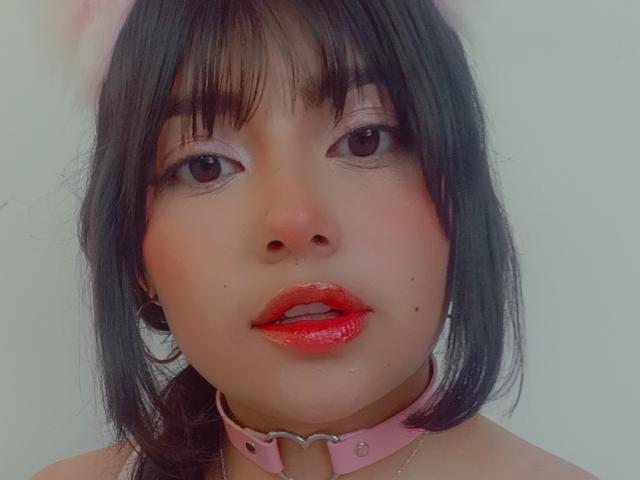 A NekoPam tem cabelo preto, olhos rosa, batom vermelho e uma coleira rosa com tachas de metal. Seu rosto está centralizado, mostrando uma expressão fofa e ligeiramente sedutora..