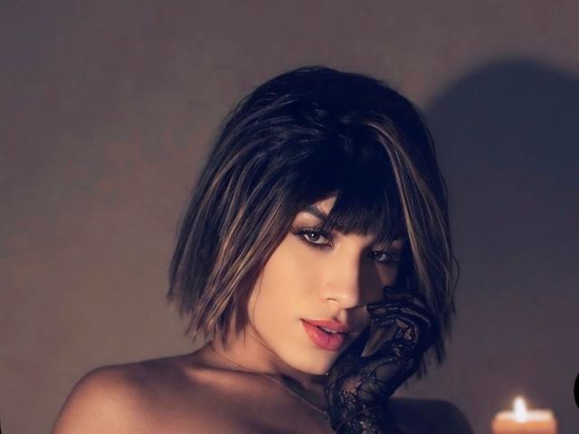 SiaraRealish avec une coupe de cheveux courte et sombre, portant un gant en dentelle noire, regardant séductrice l'appareil photo, lèvres rouges, lumière douce, bougie en arrière-plan. Les seins ne sont pas visibles, l'accent est mis sur le visage et le gant..