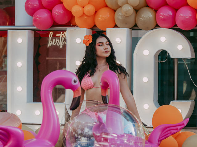 SophyCarter, avec de longs cheveux noirs, une fleur orange dans les cheveux, un maillot de bain rose, un flamant rose gonflable, des ballons orange et rose, de grandes lettres blanches avec des lumières en arrière-plan..