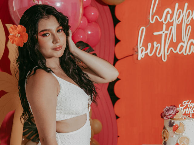 Sophy Carter, avec ses cheveux noirs ondulés, porte un crop top blanc en dentelle, a un maquillage rose pour les yeux et une fleur orange dans ses cheveux, et se tient debout avec une main sur sa tête..