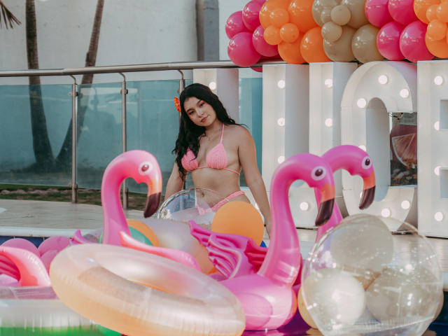 SophyCarter en maillot de bain rose, avec un bas orange, cheveux noirs, une fleur orange, agenouillée sur un flamant rose gonflable, entourée d'autres objets gonflables et de ballons, près de la piscine, sous une lumière douce..