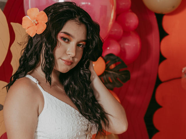 Sophy Carter, com cabelo escuro e encaracolado, uma flor laranja, uma blusa branca de renda e maquiagem vermelha nos olhos, com a mão no cabelo, olhando para a câmera, com um sorriso suave, com balões rosa e laranja ao fundo..