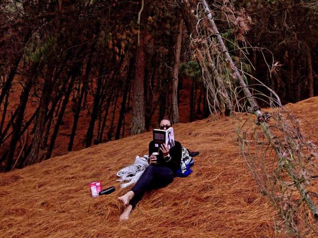 AlexiaJamesX, descalça, deitada sobre agulhas de pinheiro, lendo um livro, vestindo roupas pretas e brancas, com uma bolsa azul e uma caixa rosa perto dela, em uma floresta..