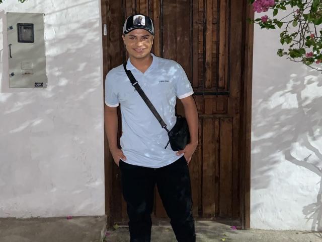 ThiagoAriass steht mit den Händen in den Taschen seiner schwarzen Hose, trägt ein weißes Polohemd, eine schwarze Kappe und eine schwarze Umhängetasche, vor einer Holztür. Er lächelt leicht..