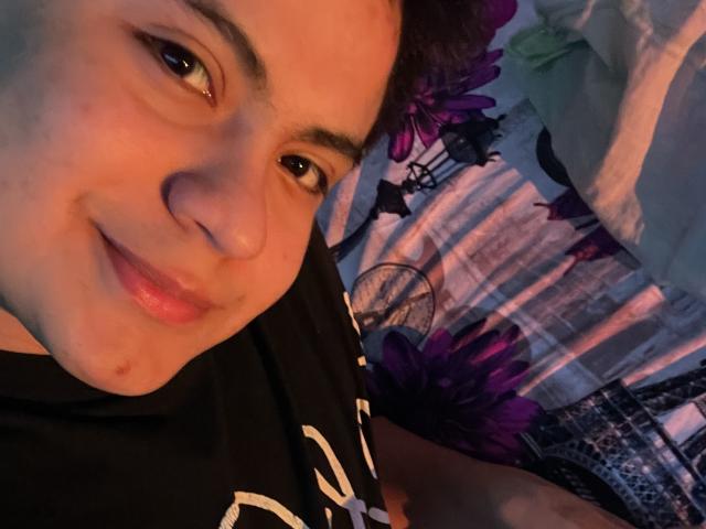 ThiagoAriass, sorridente, con occhi marrone chiaro, capelli corti viola e pelle chiara, indossa una maglietta nera, in un selfie, mostrando una piccola macchia sotto l'occhio destro..