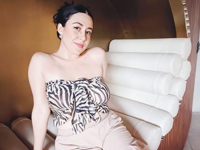 MabyLegrand met een top zonder bandjes met zebraprint, die haar grote borsten laat zien, zittend op een witte leren stoel, met beige broek, donker haar in een knot, lachend, oorbellen, lichte huid en roze lippenstift..