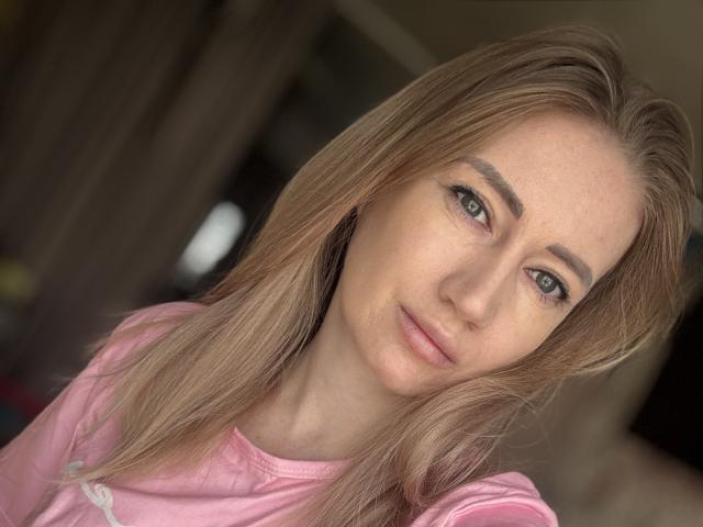 JessikaBella met blond haar, blauwe ogen en een roze shirt, kijkt in de camera met een lichte kanteling van het hoofd, een zachte uitdrukking, subtiele make-up en een natuurlijke huid. Haar borsten zijn niet zichtbaar..