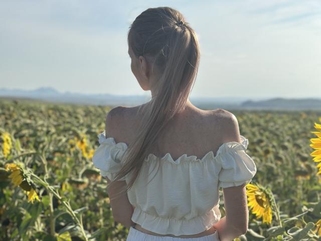 JessikaBella with long blonde ponytail, white off-shoulder top, standing in sunflower field, back to camera, sunlit, soft light, yellow flowers, green leaves, clear sky, calm atmosphere.
