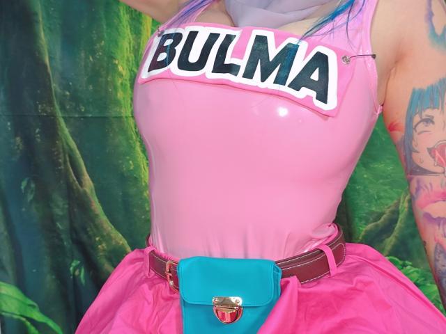 Kkole has pink latex boobs, a shiny pink skirt, a blue belt with a heart buckle, and tattoos on her arm. Her blue hair is visible.