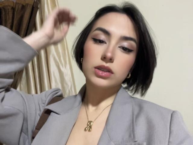 Mia Harper, con cabello negro corto, vestida con un blazer gris, un collar dorado y lápiz labial rosa, levantando su mano derecha y mirando ligeramente hacia abajo. Sus senos son parcialmente visibles..