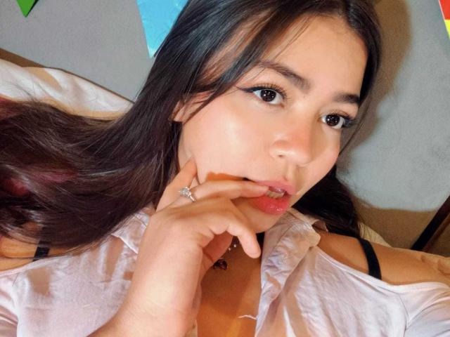 KimDevinne with long brown hair, looking at camera, sucking her finger, white shirt, black strap, ring on finger, brown eyes, subtle makeup, slightly parted pink lips, soft light.