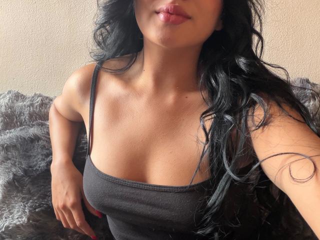 Lilly Taylor, con i capelli scuri e ondulati, labbra piene, pelle marrone, indossa una canotta nera che mette in mostra il suo seno, di dimensioni piccole o medie, ed è seduta su una superficie grigia e pelosa..