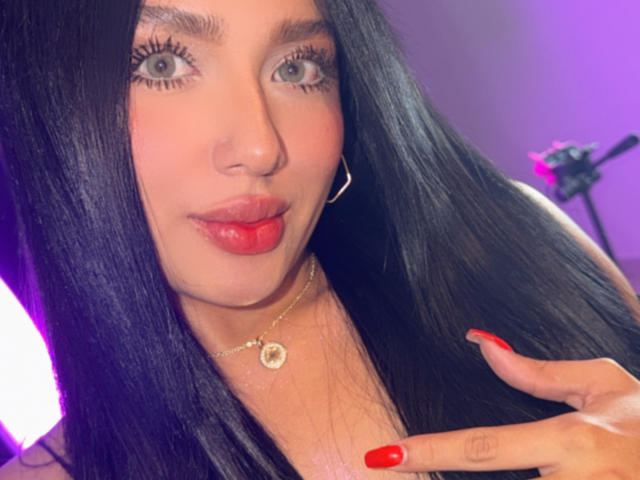 Lilly Taylor ha lunghi capelli neri, rossetto rosso e unghie rosse, indossa una collana dorata e orecchini a cerchio, e fa un gesto di "silenzio" portando il dito alle labbra..