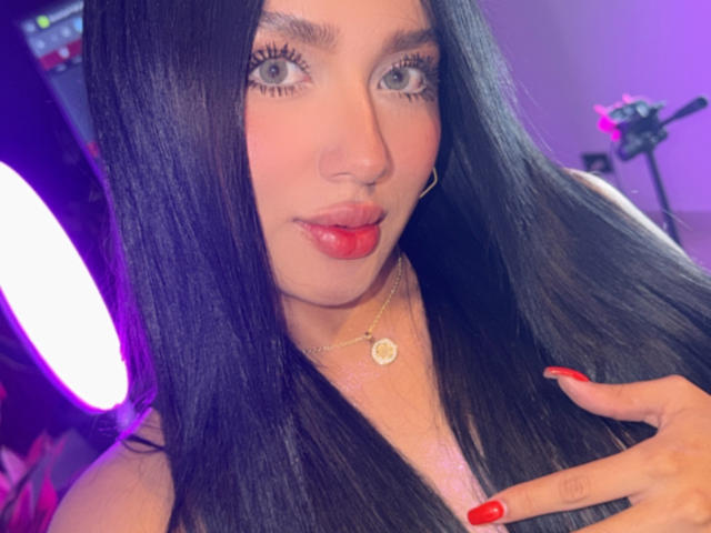 Lilly Taylor, con lunghi capelli neri, labbra rosse, occhi verdi, unghie rosse, una collana d'oro e un piercing al naso, guarda direttamente l'obiettivo. La sua mano tocca il suo petto..