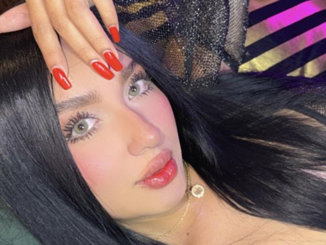 Lilly Taylor, con cabello negro brillante, ojos verdes, uñas rojas, labios rosados y un collar de oro, está acostada con la mano en la cabeza. Sus senos son parcialmente visibles, y lleva puesto un top negro de encaje..